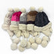Five knitted winter hats with pom-poms and ear flaps, displayed side by side in various colors. They have fluffy, cream-colored lining and attached braided tassels ending in pom-poms.