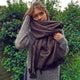 A person wrapped in a large brown scarf stands outdoors, holding it close, wearing a grey sweater. The background features lush green foliage with small pink flowers.