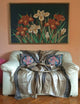 Mogato baby alpaca wool blanket – a cream armchair holds two decorative pillows and a draped beige-striped throw. Above, a framed painting of red and white flowers against a dark background adorns a salmon-colored wall