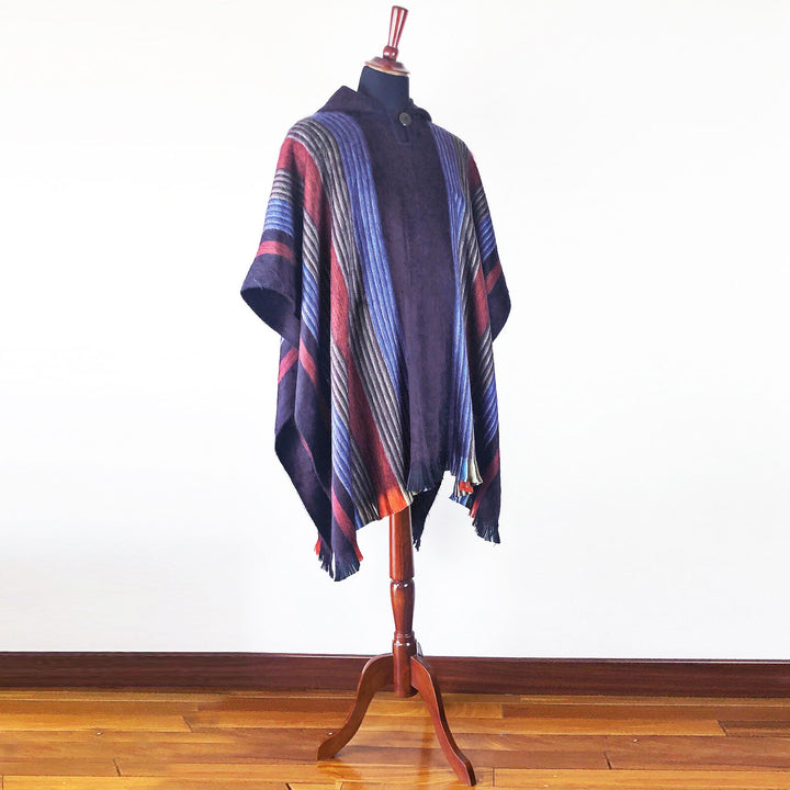 A multicolored striped poncho is displayed on a wooden mannequin stand, set against a plain white wall with a wooden floor.