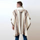 A person wears a cream poncho with brown geometric patterns, standing against a plain white background. The poncho has a hood and fringed edges.