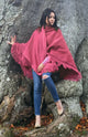 A person wrapped in a fringed pink shawl stands against a large tree, wearing blue ripped jeans and beige heels, surrounded by fallen leaves.