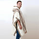 A person wears a hooded poncho with geometric patterns in brown and black on a cream background, standing sideways against a plain white backdrop.