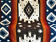 Qhusi extra large alpaca wool blanket – a woven textile displays geometric patterns with diamond shapes in white, black, and blue, accented by orange-red stripes, creating a bold, symmetrical design