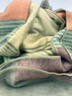Latacunga alpaca wool throw blanket – a soft, multicolored woolen blanket with pastel stripes lies loosely crumpled, showcasing its stitched edges and cozy texture in a close-up view
