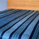 Chimborazo baby alpaca wool blanket – a bed is covered with a striped blanket featuring varying shades of blue and gray. The headboard is made of light brown wood, and the background is simple and minimalistic