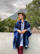 A person in a blue poncho and brown hat stands leaning on a white wall, gazing sideways. In the background, there are green plants and a mountain under a cloudy sky.