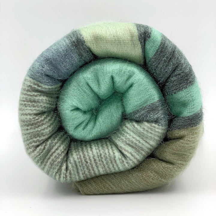 Saquisili baby alpaca wool blanket – a rolled, striped blanket with shades of green, gray, and cream is tightly coiled, resting against a plain, white background