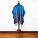 A blue textile poncho with colorful patterns and fringe drapes over a mannequin. It stands in a room with wooden flooring and a plain white wall.