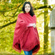 A person wrapped in a red fringed shawl stands still, wearing a pink sweater and jeans, surrounded by lush green foliage on a sunny day.
