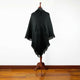 A black poncho drapes over a wooden mannequin stand, featuring a front pocket and fringed edges, set against a plain white background and wooden floor.