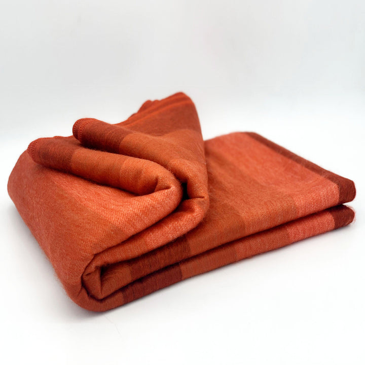 Cusubamba baby alpaca wool blanket – a neatly folded, soft orange blanket rests on a smooth, white surface, showcasing subtle shading variations and a cozy texture