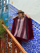 A person in a wide-brimmed hat adjusts their fringed poncho while standing near blue patterned tiles and a wooden staircase railing, with greenery in the foreground.