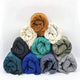 Rolled scarves, arranged in a pyramid, display various colors including teal, blue, gray, and brown, set against a plain white background.
