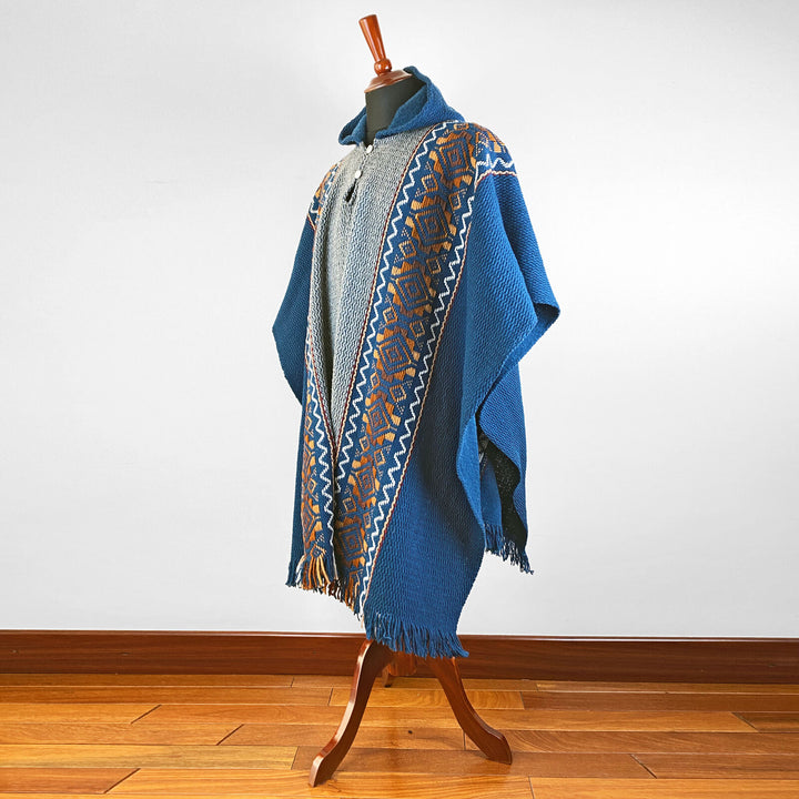 A blue poncho adorned with intricate geometric patterns is displayed on a wooden mannequin stand, set against a plain indoor background with wooden flooring.