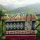 A colorful patterned blanket hangs on a black metal railing, displaying geometric designs. A potted plant with pink flowers is on the left, with a mountainous landscape in the background.
