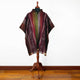 A colorful poncho displayed on a wooden mannequin stand features intricate patterns and vertical stripes. It stands in a room with hardwood flooring and a plain white wall as the backdrop.