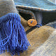 A soft, multi-colored woolen fabric features a brown button and vibrant blue fringe, set in a cozy, woven texture.