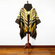 A multicolored poncho with geometric patterns hangs on a wooden stand, displayed against a white wall and hardwood floor.