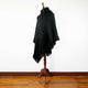 A black poncho with a fringe hem and hood is draped over a wooden mannequin stand placed on a polished wooden floor against a plain white wall.