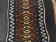 A woven textile features intricate geometric patterns in brown, orange, and black, accented by white zigzag lines, creating a vibrant and textured design.