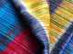 Alpaca wool queen blanket – a colorful knitted fabric features a repeated stripe pattern in blue, red, yellow, and purple, gently folded. The texture appears soft and fuzzy in a close-up view