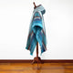 A multicolored striped poncho with a hood hangs on a wooden stand, displayed against a plain white wall on a wooden floor.