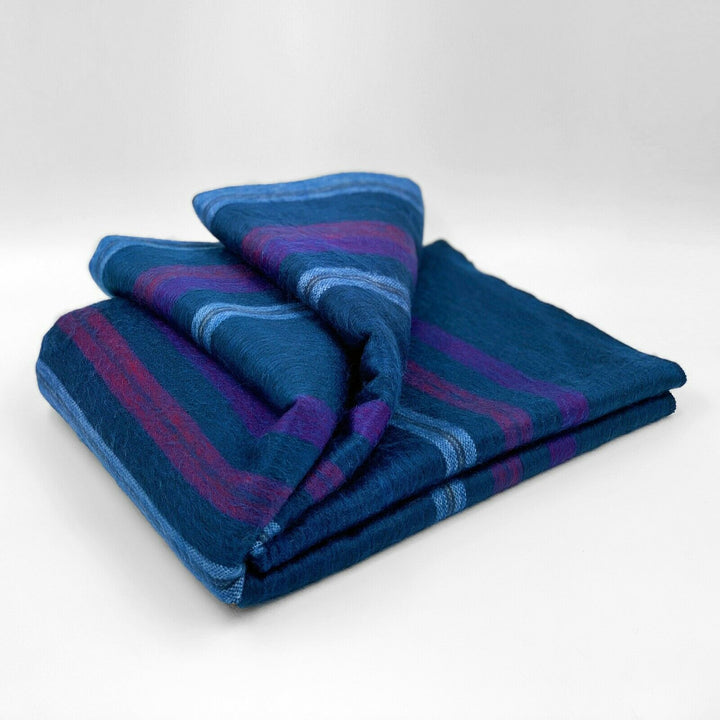 Tamboloma baby alpaca wool blanket – a folded, striped blanket in various shades of blue and purple rests on a smooth, white surface, displaying its soft texture and vibrant colors