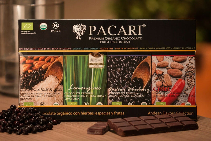 A package of Pacari Premium Organic Chocolate featuring four flavors: Cuzco Pink Salt & Nibs, Lemongrass, Andean Blueberry, and Chile. It is labeled as fine, organic Ecuadorian chocolate. Several certifications and awards are depicted. Some chocolate squares and berries are positioned in front of the package.