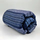 Chimborazo baby alpaca wool blanket – a rolled-up, blue and gray striped blanket rests on a smooth, white surface, suggesting a cozy and inviting context