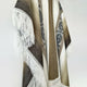 A patterned wool blanket hangs with white fringe edges, displaying geometric designs in earthy tones against a plain white background.