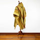 A wool poncho drapes over a wooden stand, featuring geometric patterns and warm colors, set against a plain white background and wooden floor.