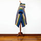 A colorful poncho with geometric patterns hangs on a wooden mannequin. It has shades of blue, green, and beige, and is displayed against a plain white wall and wooden floor.
