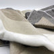 Mogato baby alpaca wool blanket – a soft, multi-toned woolen blanket lays draped and overlapping, displaying shades of gray, beige, and white with visible stitching, set against a plain, light background