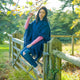 A person wearing a blue shawl and ripped jeans sits on a wooden gate, holding a tree branch, surrounded by a lush, sunny green landscape.