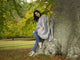 A person sits against a large tree, wrapped in a gray shawl, surrounded by lush green leaves in a serene park setting, with autumn leaves scattered on the ground.