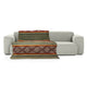 A patterned blanket drapes over a simple, light gray sofa. The blanket features geometric diamond designs in red and brown hues against an olive and beige background, creating a cozy atmosphere.