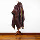 A woven poncho with maroon, brown, and green patterns drapes over a wooden mannequin. It's situated in a room with wooden flooring and a plain white wall.