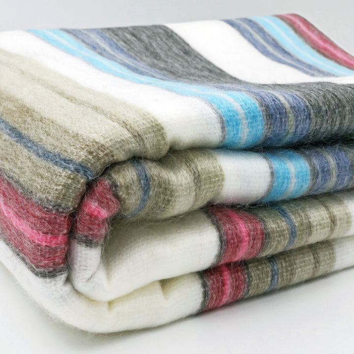 Carihuairazo baby alpaca wool blanket – a folded woolen blanket with wide horizontal stripes in white, gray, blue, and pink rests neatly on a light surface, suggesting warmth and comfort in a minimalist setting