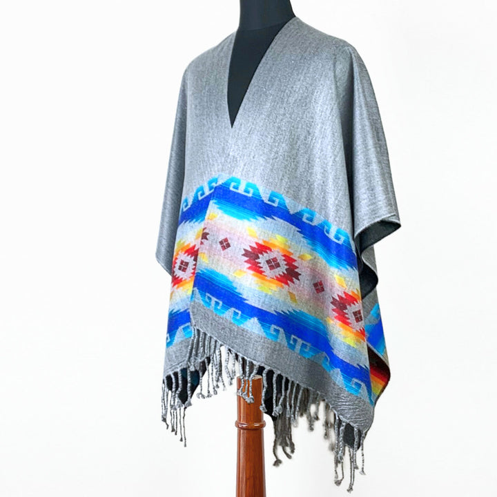 A gray poncho with colorful geometric patterns hangs from a mannequin in a well-lit studio. Fringes dangle from the bottom edge, adding detail to the garment.