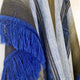 A woven textile hangs vertically, showcasing intricate patterns in shades of blue and brown, with prominent blue fringes. The fabric's texture and design add depth against a plain background.