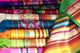 Colorful woven textiles are stacked and arranged, displaying intricate patterns. The fabrics include stripes, geometric designs, and fringed edges, highlighting a vibrant assortment of colors in a display setting.