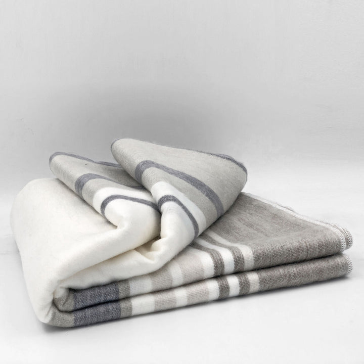 Yacoubina baby alpaca wool blanket – a folded blanket with gray and white stripes rests on a flat, light surface, exhibiting a soft and cozy texture in a minimalistic setting