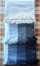 Three fringed woolen scarves, stacked vertically in an overlapping pattern, display shades of cream, gray, and blue. They rest on a checkered, brown and beige tablecloth background.
