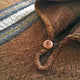 A wooden button fastens a loop on a brown, textured cloth, featuring multicolored horizontal stripes of blue, green, and white, suggesting a cozy garment design.