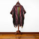 A hooded poncho with geometric patterns hangs on a wooden stand, placed on a polished wooden floor against a plain white wall.