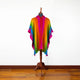 A vibrant, multicolored poncho drapes over a wooden mannequin stand, set against a plain white wall. The poncho features bold stripes in pink, orange, green, and blue hues.