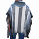 A person wears a striped poncho with geometric patterns, standing with their back to the viewer. The poncho features a hood and is set against a plain background.