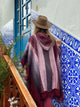A person wearing a hat and poncho walks up stairs beside a blue-tiled wall with floral patterns, leading toward a blue door. Plant branches are visible along the railing.