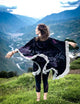 A person stands with arms outstretched, wearing a fringed shawl, overlooking a lush, mountainous landscape with a winding river and distant buildings.
